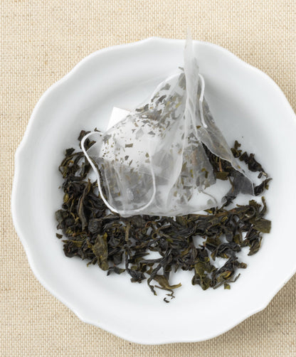 A pyramid-shaped tea sachet with loose leaves scattered around it on a white plate, set against a beige fabric background.