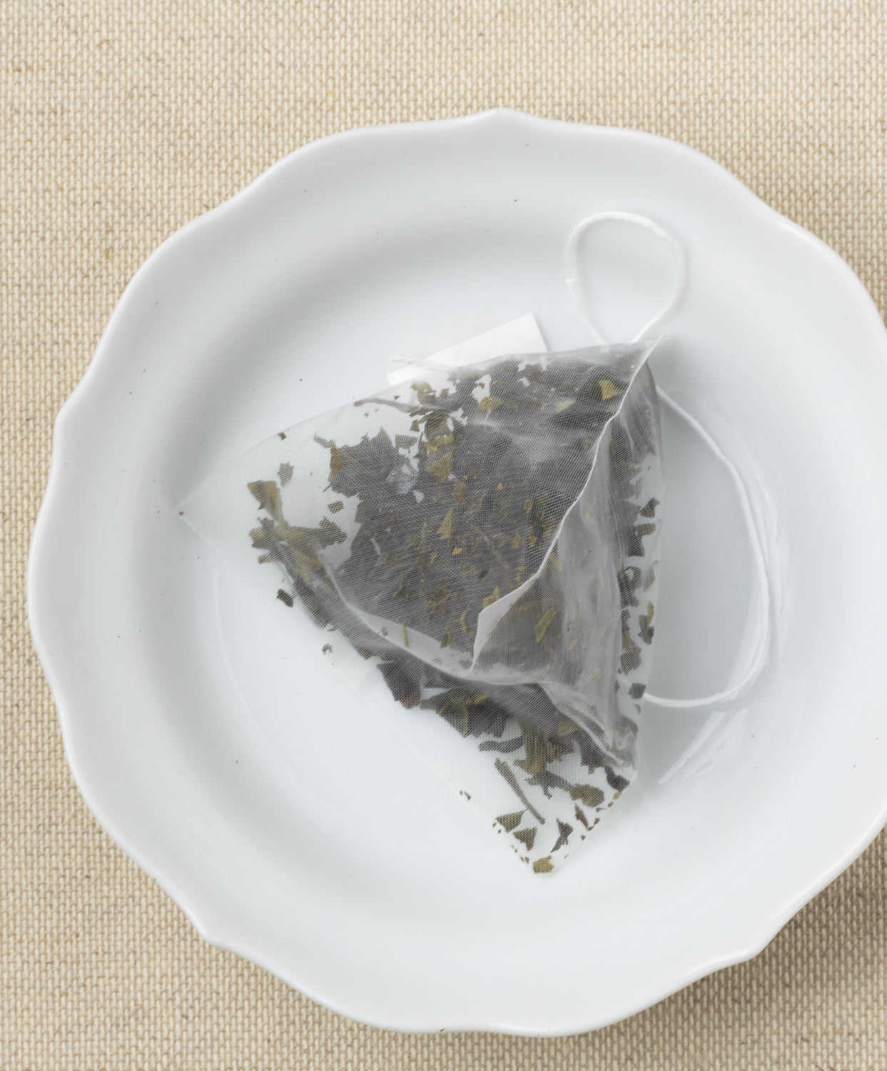 A pyramid-shaped tea sachet filled with dried leaves, displayed on a white plate against a beige background.