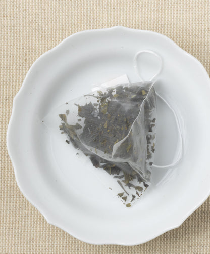 A pyramid-shaped tea sachet filled with dried leaves, displayed on a white plate against a beige background.